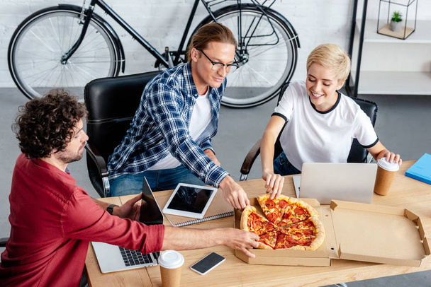 high angle view of smiling young coworkers eating pizza at workplace  - Photo, Image