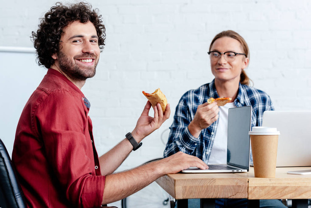 giovani uomini sorridenti mangiare pizza e utilizzando computer portatili in ufficio
 - Foto, immagini