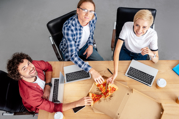 korkea kulma näkymä onnellisia nuoria kollegoja syö pizzaa ja hymyilee kameralle työpaikalla
 - Valokuva, kuva