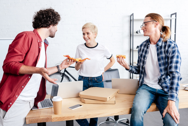 happy young colleagues holding pizza and smiling each other in office  - 写真・画像