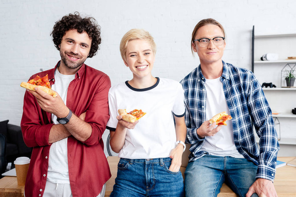 szczęśliwy młody uruchamiania team gospodarstwa pizza i uśmiecha się do kamery w biurze - Zdjęcie, obraz