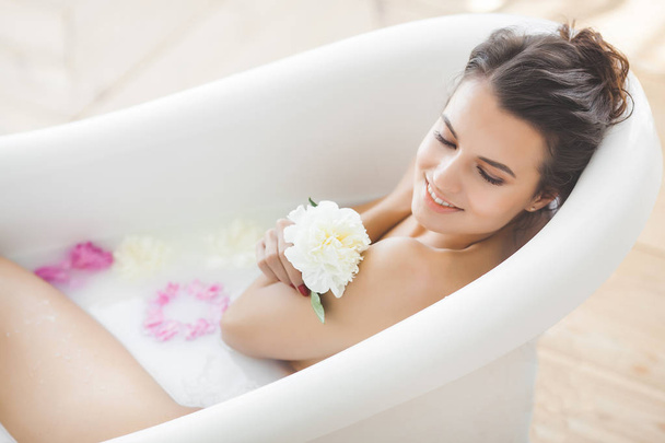 Νεαρή όμορφη γυναίκα που κάνει μπάνιο με λουλούδια και γάλα - Φωτογραφία, εικόνα