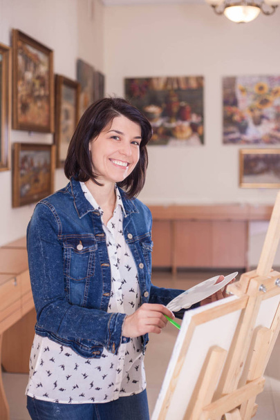 Ładna, młoda kobieta, malarstwo obraz w pobliżu sztalugach w creative studio. - Zdjęcie, obraz