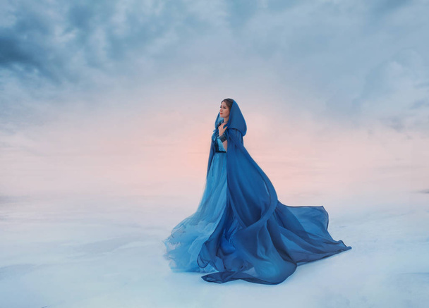 The Snow Queen in a blue raincoat that flutters in the wind. A traveler on a background of sunrise or sunset, and a frozen valley covered with snow and clouds, a place where heaven meets the earth - Foto, Bild