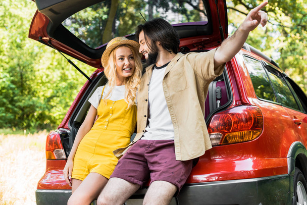 boyfriend pointing on something to girlfriend while sitting on car trunk - Фото, зображення