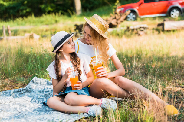 mother and daughter sitting on blanket at picnic, looking at each other and holding glasses of juice - Fotoğraf, Görsel