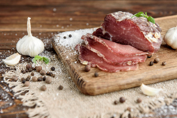Χοιρινό αποξηραμένο κρέας φέτες σε ρουστίκ σκούρο ξύλινο φόντο. Αποξηραμένο χοιρινό προσούτο σαλάμι με βότανα. - Φωτογραφία, εικόνα
