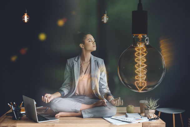žárovka představující kreativní myšlení a mladá podnikatelka meditoval v lotosové pozici na pracovním stole - Fotografie, Obrázek