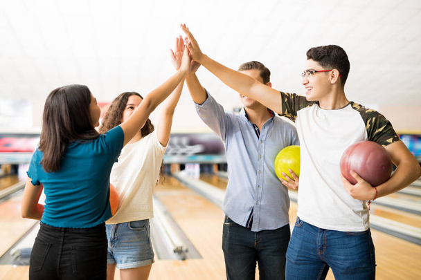 Male and female teenage friends giving high-five while holding bowling balls at alley in club - Photo, Image