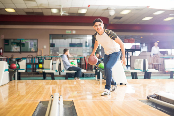 Full length of smiling teenage boy throwing ball while practicing bowling game in club - Foto, Bild