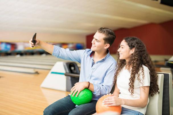 Sonriente pareja adolescente con bolas de bolos tomando autorretrato mientras usa el teléfono inteligente en el club
 - Foto, Imagen