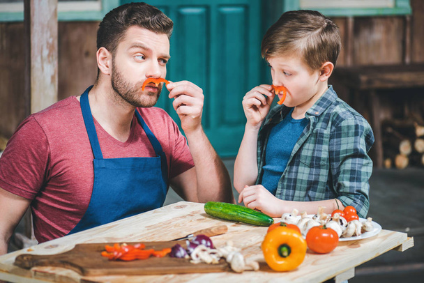 маленький мальчик с отцом делает усы с ломтиками перца во время приготовления пищи за столом
  - Фото, изображение