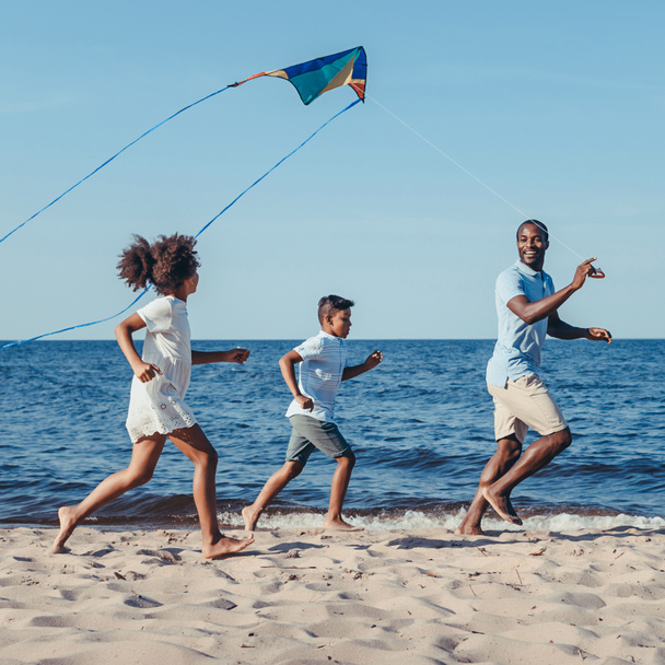 side view of happy african american father and kids playing with kite on beach - Photo, Image