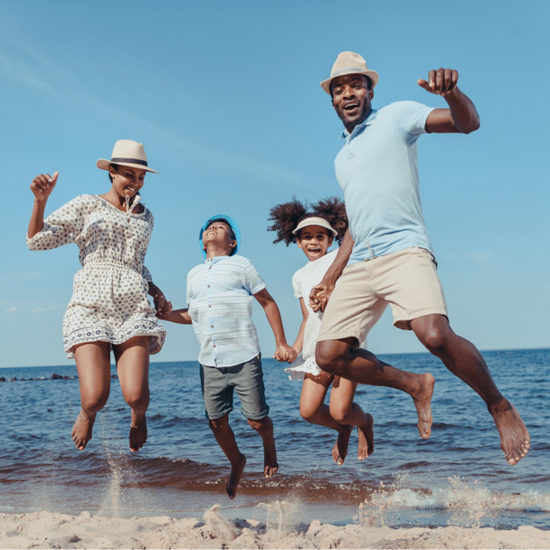 молодая счастливая африканская американская семья держась за руки и прыгая на пляже в солнечный день
 - Фото, изображение