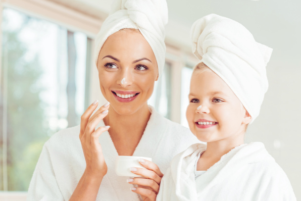 attraktive lächelnde Mutter und Tochter in Bademänteln und Handtüchern, die gemeinsam Gesichtscreme auftragen - Foto, Bild