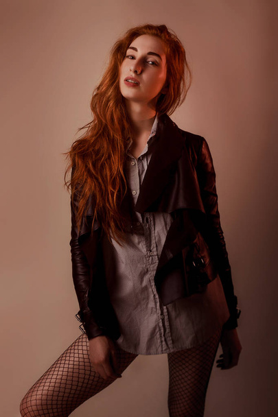 Fashion studio portrait of woman with red long hair in shirts and leather jacket and black fishnet tights, warm tones, fashion and style concept - 写真・画像