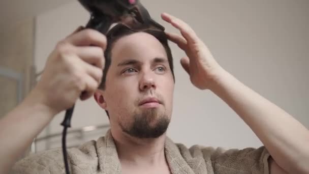 Portret van een jonge man die droogt natte haren met behulp van een elektrische haardroger - Video