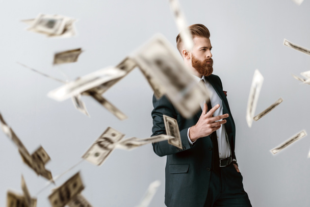 handsome elegant businessman with falling dollar banknotes on foreground isolated on grey - Photo, Image