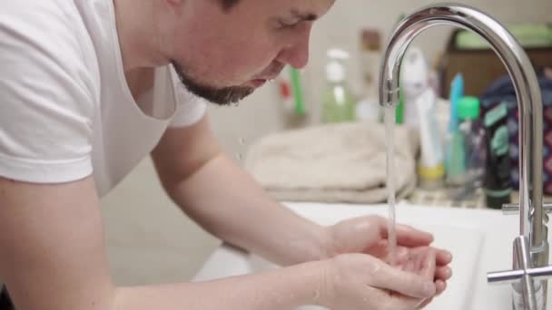 Egy fiatal és szakállas ember mossa az arcát a csap a fürdőszobában alatt - Felvétel, videó