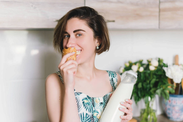 Smiling woman holding bread and milk bottle portrait on kitchen. Woman in the kitchen eating baguette. Lifestyle - Zdjęcie, obraz