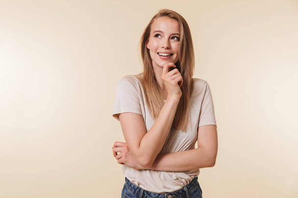 Image of affable blond woman 20s wearing casual t-shirt smiling and looking upward while standing with arms crossed isolated over beige background in studio - Fotoğraf, Görsel