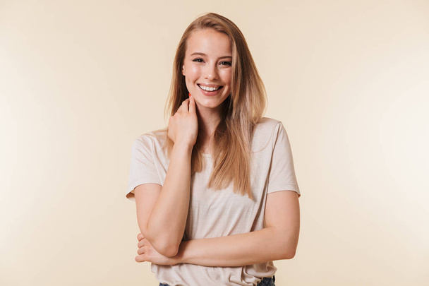 Portrait of charming blonde woman 20s wearing casual t-shirt smiling at camera while touching neck isolated over beige background in studio - Фото, изображение