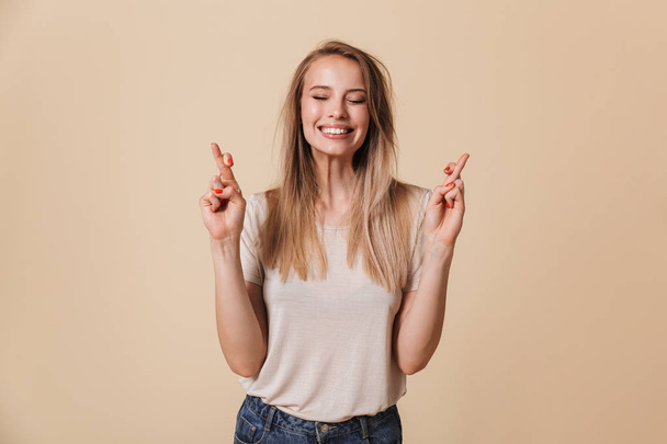 Portrait of a smiling casual girl holding fingers crossed for good luck isolated over beige background - Foto, afbeelding