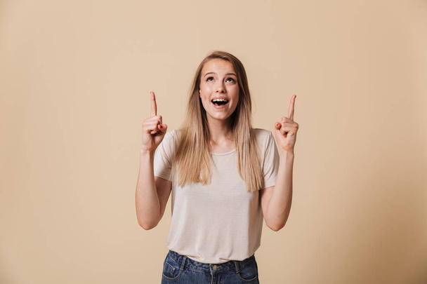 Portrait of a happy casual girl pointing finger up at copy space isolated over beige background - Fotoğraf, Görsel
