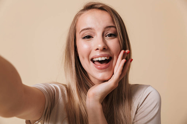 Portrait of a cheerful young girl taking a selfie with outstretched hands isolated over beige background - Photo, Image
