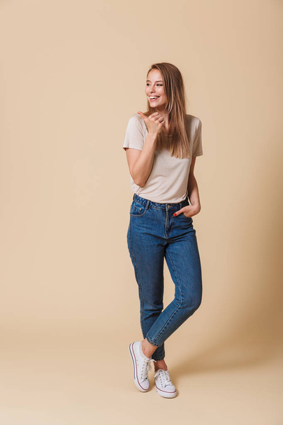 Full length portrait of a smiling casual girl pointing finger away at copy space isolated over beige background - Fotografie, Obrázek