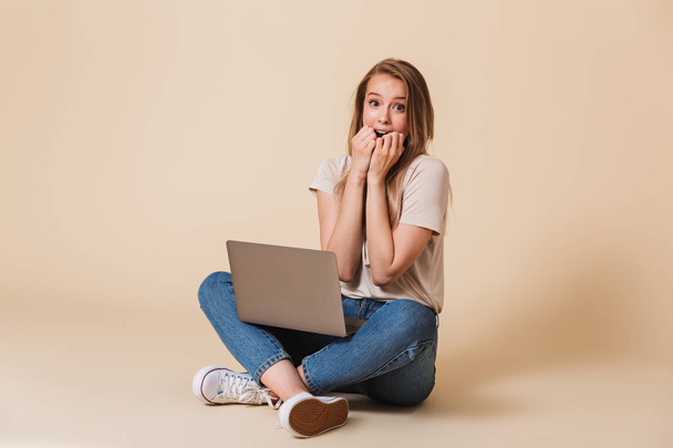 Portrait of an excited casual girl with laptop computer sitting on a floor with legs crossed isolated over beige background - Fotoğraf, Görsel