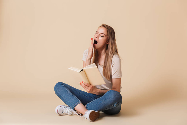 Photo of bored tired woman wearing casual clothing sitting on floor with legs crossed and yawning while reading uninteresting book isolated over beige background - Photo, image