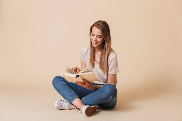 Photo of pretty young woman wearing casual clothing smiing while sitting on floor with legs crossed and reading interesting book isolated over beige background - Foto, Bild