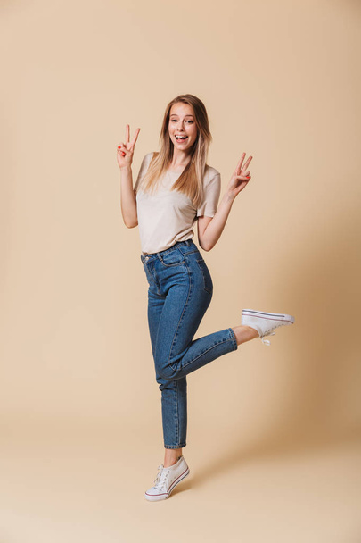 Full length portrait of satisfied young woman 20s wearing casual clothing smiling and showing peace sign with two hands isolated over beige background - Фото, изображение