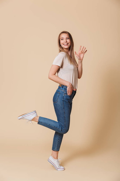 Full length portrait of attractive blond woman 20s wearing casual clothing lifting her leg and smiling while waving hand isolated over beige background - Fotoğraf, Görsel