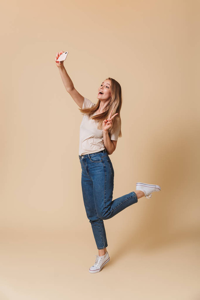Full length photo of positive blond woman 20s wearing casual clothing taking selfie photo on cell phone isolated over beige background - Photo, Image