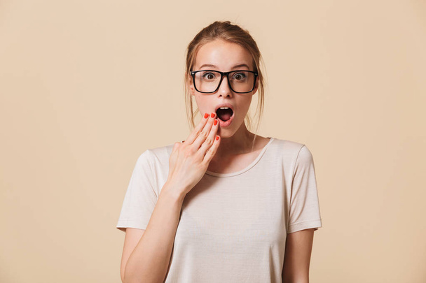 Image of surprised blonde woman 20s with tied hair wearing basic t-shirt and eyeglasses covering open mouth with hands isolated over beige background in studio - Фото, зображення