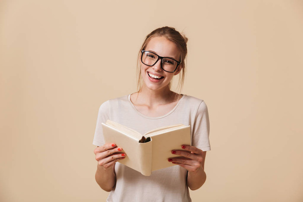 Portrait of a lovely young girl in eyewear reading a book isolated over beige background - Fotoğraf, Görsel