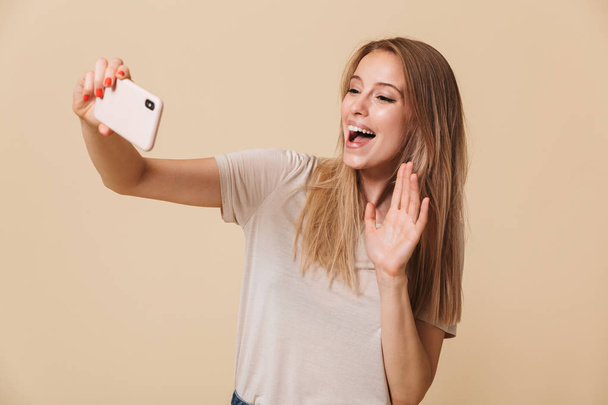 Portrait of a pretty young girl taking a selfie and waving hand isolated over beige background - Foto, afbeelding