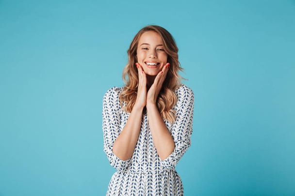 Cheerful blonde woman in dress touching her cheeks and looking at the camera over blue background - Photo, image
