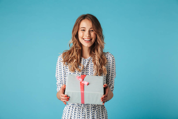 Happy blonde woman in dress holding gift box and looking at the camera while enjoys over blue background - Foto, immagini