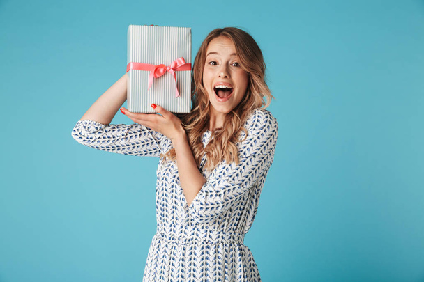 Shocked blonde woman in dress holding gift box while looking at the camera over blue background - Foto, Bild
