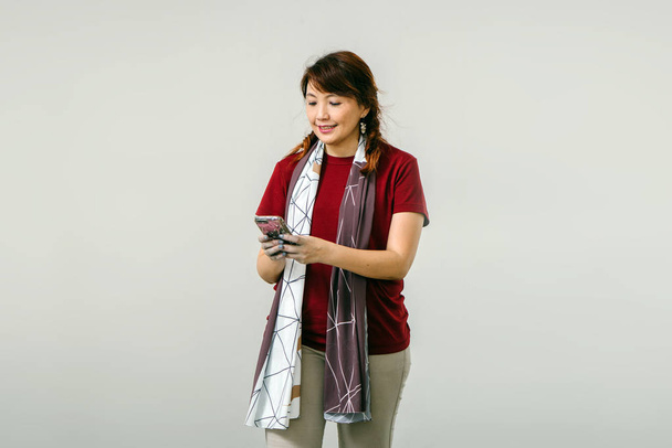asian woman wearing red blouse smiling texting on mobile phone standing on white studio background - Photo, Image