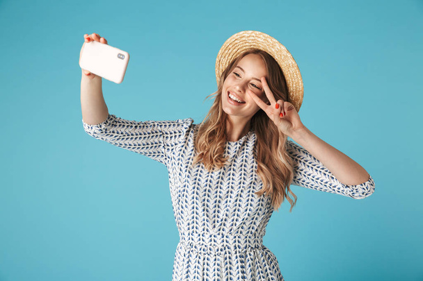 Smiling blonde woman in dress and hat making selfie on smartphone while showing peace gesture over blue background - Photo, Image