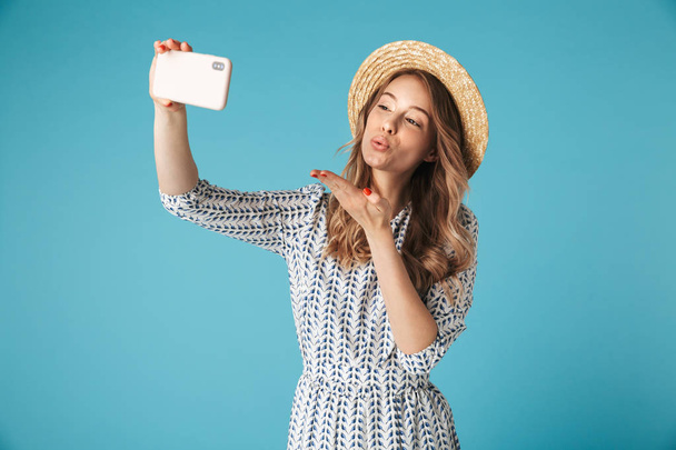 Happy blonde woman in dress and hat making selfie on smartphone while sends air kiss over blue bckground - Zdjęcie, obraz