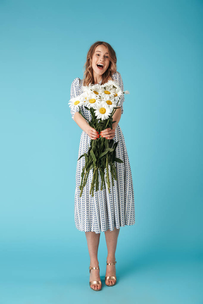 Full length image of Cheerful blonde woman in dress giving flowers at camera over blue background - Zdjęcie, obraz