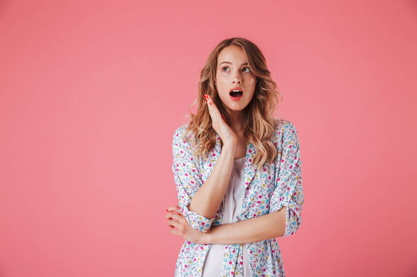 Portrait of a surprised young woman in summer dress looking away at copy space isolated over pink background - Foto, afbeelding