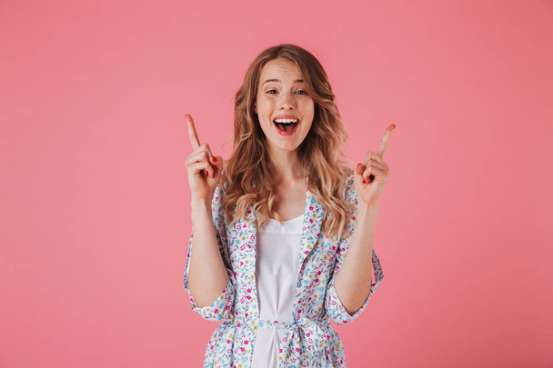 Portrait of an excited young woman in summer dress pointing finger up at copy space isolated over pink background - Foto, imagen