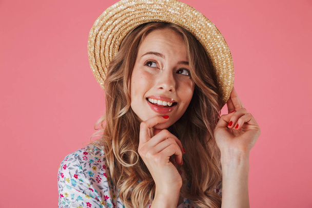 Close up portrait of a smiling young woman in summer dress and straw hat looking away isolated over pink background - Photo, image