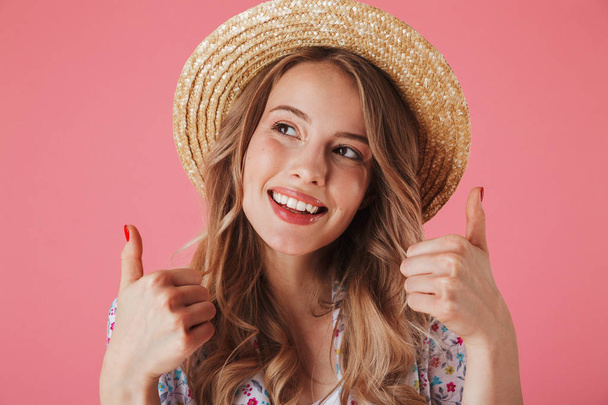 Close up portrait of a happy young woman in summer dress and straw hat showing thumbs up gesture and looking away isolated over pink background - Fotografie, Obrázek